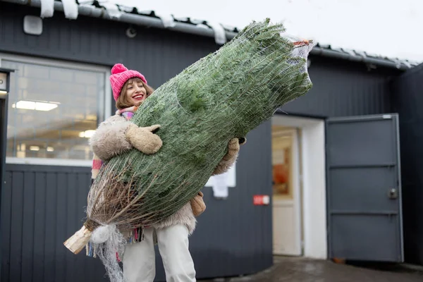 stock image Young woman carries wrapped Christmas tree while going out from post office delivered from online shop. Concept of delivery and online shopping on winter holidays