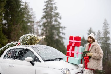 Kadın, Noel çelengi ve çatıda ağaçla süslenmiş arabanın yanında dikilmiş, karlı havada dağlarda arabayla seyahat ederken bir yığın hediye kutusu tutuyor. Kış tatili kavramı
