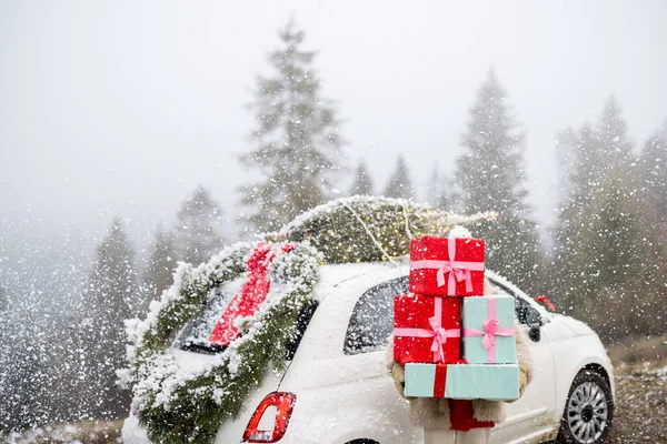 女性は雪の日に山の中を車で移動しながら 屋上のクリスマスリースと木で飾られた車のそばに立っている間 ギフトボックスのヒープを保持しています 冬休みの概念 — ストック写真