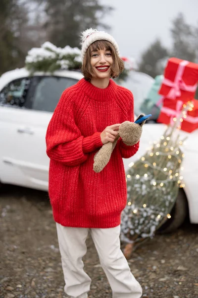 身穿红色毛衣的女人站在圣诞树旁 拿着手机 在山里开车送礼物 准备愉快的寒假的概念 — 图库照片