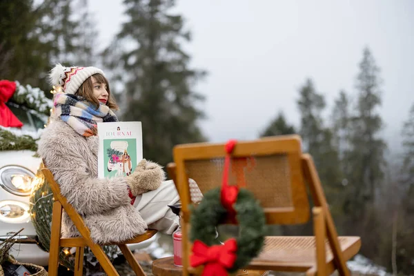 Young woman relaxes and enjoys calm on nature during winter holidays, sitting at picnic decorated for Christmas with a magazine in mountains