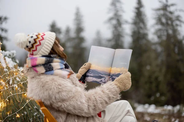 Young woman relaxes and enjoys calm on nature during winter holidays, has a picnic and reads some magazine while sitting near lighted Christmas tree in mountains