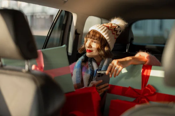 身着节日服装的年轻女子坐在后座上开车 带着包装精美的圣诞礼物和电话 为迎接寒假 准备及购物概念 — 图库照片