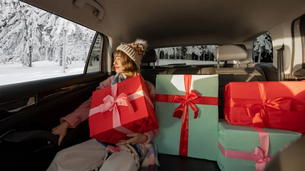 身着节日服装的年轻女子坐在后座上开车 带着包装精美的圣诞礼物 背景上的雪景 广角镜头 — 图库照片