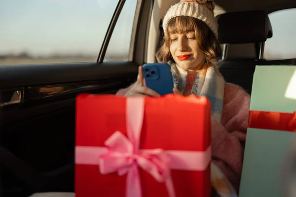 美しいラップクリスマスプレゼントと携帯電話を使用して後部座席の車の中で休日のドライブのために身を包んだ若い女性 冬の休暇を見越して 準備とショッピングのコンセプト — ストック写真
