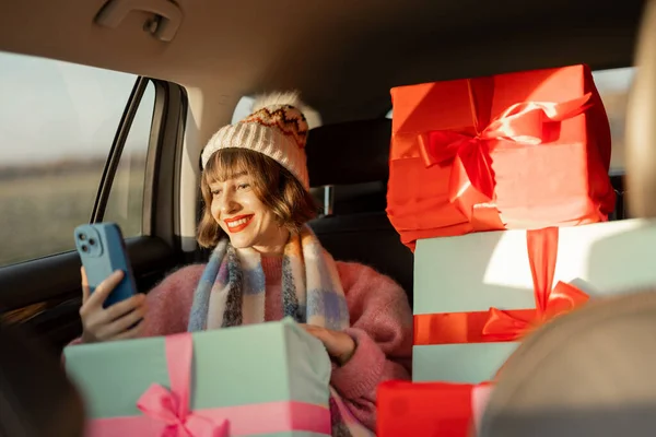 身着节日服装的年轻女子坐在后座上开车 带着包装精美的圣诞礼物和电话 为迎接寒假 准备及购物概念 — 图库照片