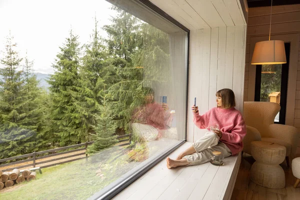 Kadın Pencere Pervazında Telefonla Oturur Doğadaki Ahşap Evde Dinlenirken Dağların — Stok fotoğraf