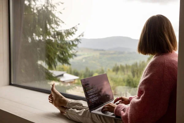 Woman works on laptop while sitting on a window with great view on mountains. Remote work from cozy place and escaping to nature concept