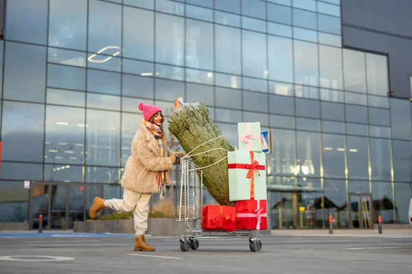 Woman pushing shopping trolley full of presents and Christmas tree in front of a mall outdoors. Winter holidays shopping concept