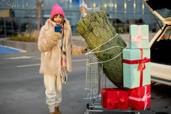 Young cheerful woman talking on phone while standing with shopping trolley full of present and Christmas tree near car at parking lot of a mall