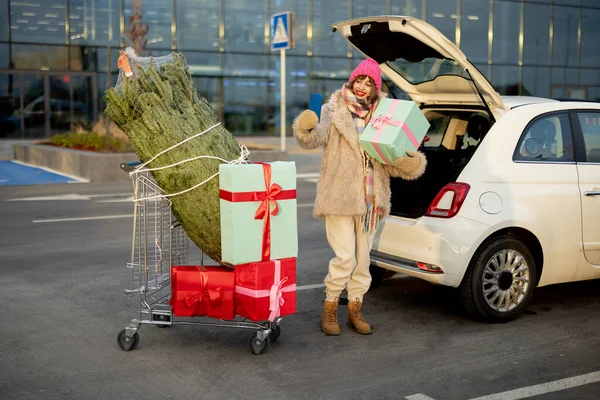 Young cheerful woman talking on phone while standing with shopping trolley full of present and Christmas tree near car at parking lot of a mall