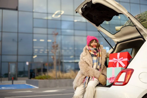 Young cheerful woman sits at car trunk full of presents near mall outdoors. Concept of winter holidays and shopping