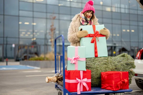 一个穿着冬衣的年轻女子站在户外购物中心附近的购物车里 满载着礼物和圣诞树的画像 冬季假日购物概念 — 图库照片