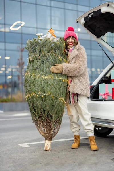 若い陽気な女性は プレゼントでいっぱいの車のトランクの近くのガーランドで飾られた包まれたクリスマスツリーで立っています モールでの冬の休日の買い物を持っています — ストック写真