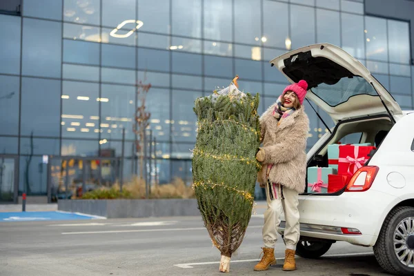 年轻而快乐的女人站在满是礼物的汽车行李箱旁 用包裹着的圣诞树装饰着花环 在购物中心过冬购物 — 图库照片