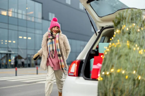 身穿冬衣的年轻而快乐的女人开着一辆满载着礼物和圣诞树的汽车在购物中心附近奔跑 — 图库照片