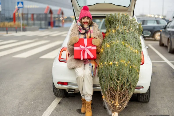 一个带着礼品盒的年轻而快乐的女人的画像 她刚买了一棵圣诞树 坐在商场停车场的汽车行李箱旁 寒假购物的概念 — 图库照片