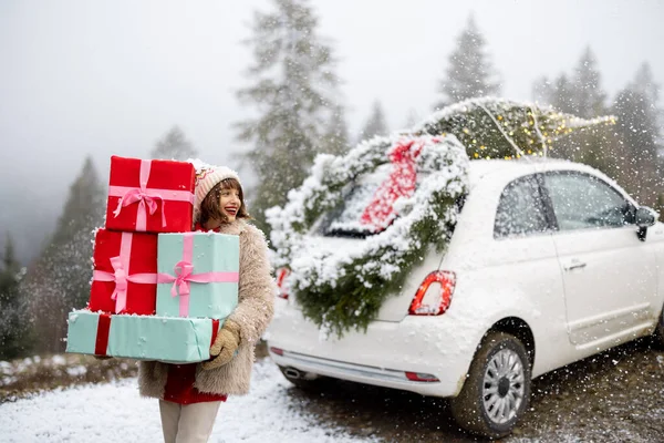 女人站在车旁 手里拿着一堆礼品盒 车顶装饰着圣诞花环和圣诞树 在多雪的天气里开车在山上旅行 寒假的概念 — 图库照片