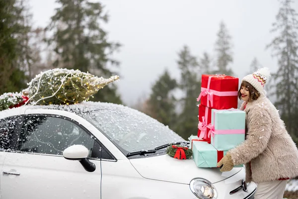 Woman holds a heap of gift boxes while standing by the car decorated with Christmas wreath and tree on a rooftop, traveling by car in the mountains on snowy weather. Concept of winter holidays