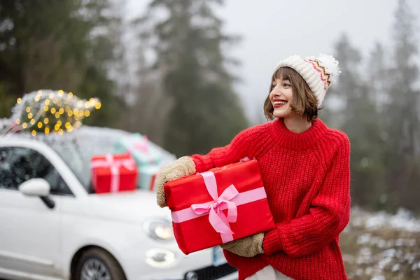 身穿红毛衣 头戴红帽子的快乐女人的画像 站在礼品盒前 坐着装饰着礼物和圣诞树的小车在山中穿梭 寒假快乐的概念 — 图库照片