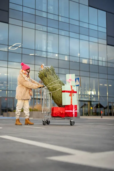 Woman pushing shopping trolley full of presents and Christmas tree in front of a mall outdoors. Winter holidays shopping concept