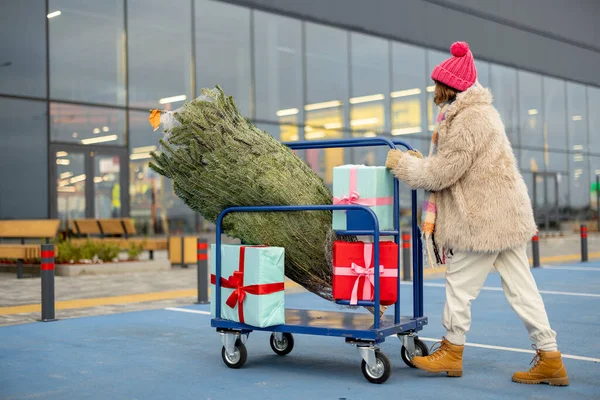 Woman pushing shopping trolley full of presents and Christmas tree at parking lot of a mall outdoors. Winter holidays shopping concept