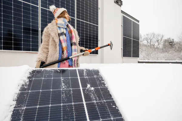 Woman cleans solar panels from snow to produce power in winter on the roof of her house on nature. Energy independence and sustainability concept. Wide angle view