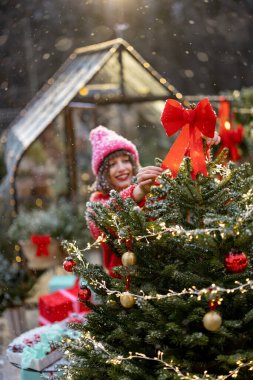 Kırmızılı genç mutlu kadın, kış tatiline hazırlanırken evinin arka bahçesinde çelenkle aydınlatılmış şenlikli toplar ve yaylarla Noel ağacını süslüyor.