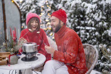 Erkek ve kadın fondüyle romantik bir akşam yemeği yerken, aynı zamanda güzel dekore edilmiş karlı arka bahçede masanın başında oturuyorlar. Genç bir aile kış tatilini dışarıda kutluyor.