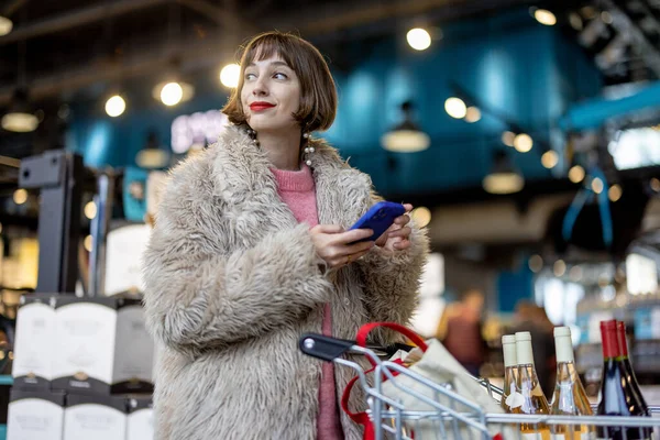 Woman with smart phone stands with a shopping cart full of wine, waiting for payment in the checkout line in supermarket