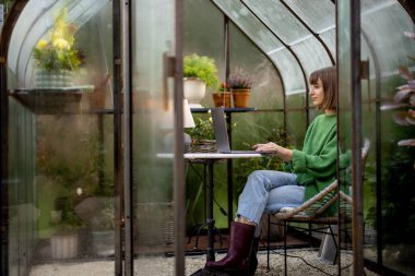 Genç bir kadın, arka bahçesinde çiçekler ve bitkilerle birlikte cam evde yuvarlak masa başında otururken dizüstü bilgisayarla çalışıyor. Doğa üzerine rahat bir atmosferde evden çalışmak