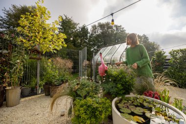 Yeşil bitkileri olan güzel bir ev, kasede küçük bir çeşme ve güneşli bir sabahta arka planda bahçeyle uğraşan bir kadın.