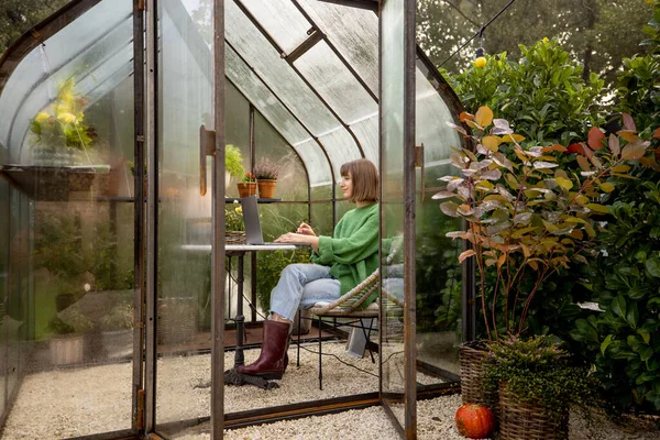 Young woman works on laptop while sitting by the round table in glasshouse with plants and flowers at backyard. Work from home at cozy atmosphere on nature