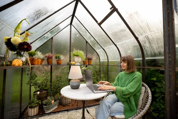 Young woman works on laptop while sitting by the round table in glasshouse with plants and flowers at backyard. Work from home at cozy atmosphere on nature