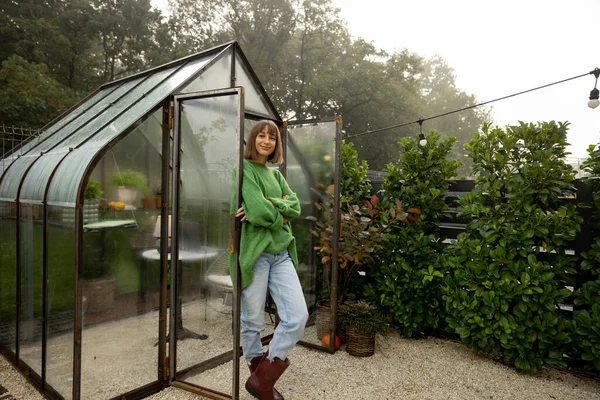 Portrait of young woman stands near greenhouse for growing plants at backyard. Gardening, leisure time in garden concept