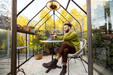 Adam arka bahçesinde çiçekler ve bitkilerle birlikte cam evde yuvarlak masa başında otururken dizüstü bilgisayarla çalışıyor. Doğa üzerine rahat bir atmosferde evden çalışmak