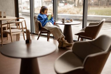 Stylish woman works on a digital tablet while sitting by the window at modern coffee shop. Concept of remote creative work online