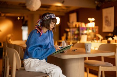 Young adult woman works on a digital tablet while sitting at modern coffee shop. Concept of remote creative work online