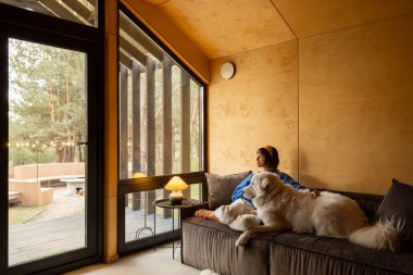Woman cares her dog while sitting on a couch by the window, spending leisure time together in wooden house on nature