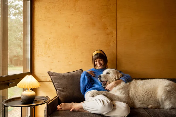 Woman cares her dog while sitting on a couch by the window, spending leisure time together in wooden house on nature