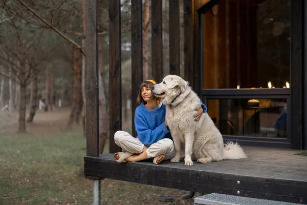 Young woman sits with her dog on porch of a wooden house in pine forest, enjoying nature while resting in cottage at countryside