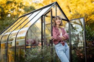Doğanın arka bahçesinde bitki yetiştirmek için güzel, kaliteli bir cam evin yanında duran neşeli bir kadın çiçekçinin portresi.