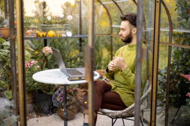 Adam dizüstü bilgisayarda çalışıyor, arka bahçesinde çiçekler ve bitkilerle cam evde yuvarlak masa başında otururken internetten konuşuyor. Doğa üzerine rahat bir atmosferde evden çalışmak