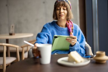 Young stylish woman works on digital tablet while sitting relaxed with coffee and cake at coffee shop. Concept of remote creative work from public place