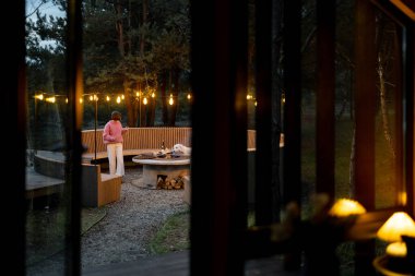 Beautiful lounge area with round bench and bonfire illuminated with garlands near house in pine forest, view from the house