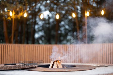 Beautiful lounge area with fireplace, round bench and illuminated garlands in forest at dusk