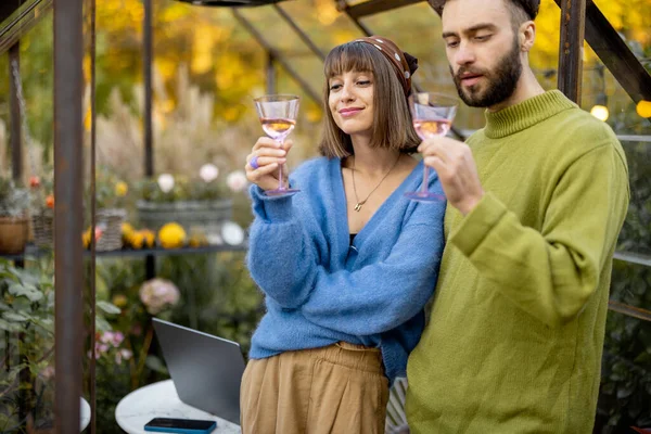 Young stylish couple talk and drink wine, spending leisure time together near glass house in garden. Man and woman have romantic eve time with alcohol drink at backyard