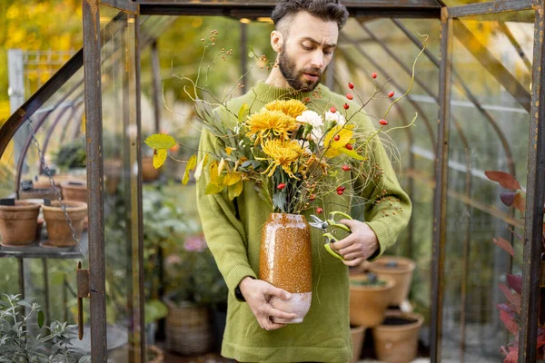 Portrait of a man florist which stands with a beautiful bouquet of flowers in front of tiny greenhouse in garden. Concept of floristry and flower workshop