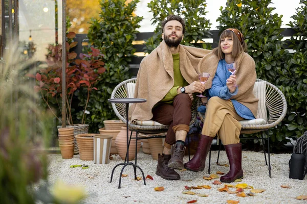 Young stylish couple warming up together, sitting covered with plaid by the fire and drink wine, spending evening time at cozy atmosphere in beautiful garden
