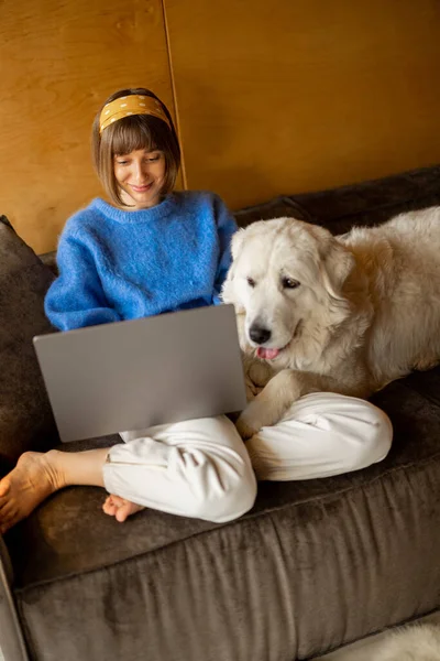 Young woman works on laptop while sitting on a couch with her white adorable dog at home. Concept of remote work from home and friendship with pets